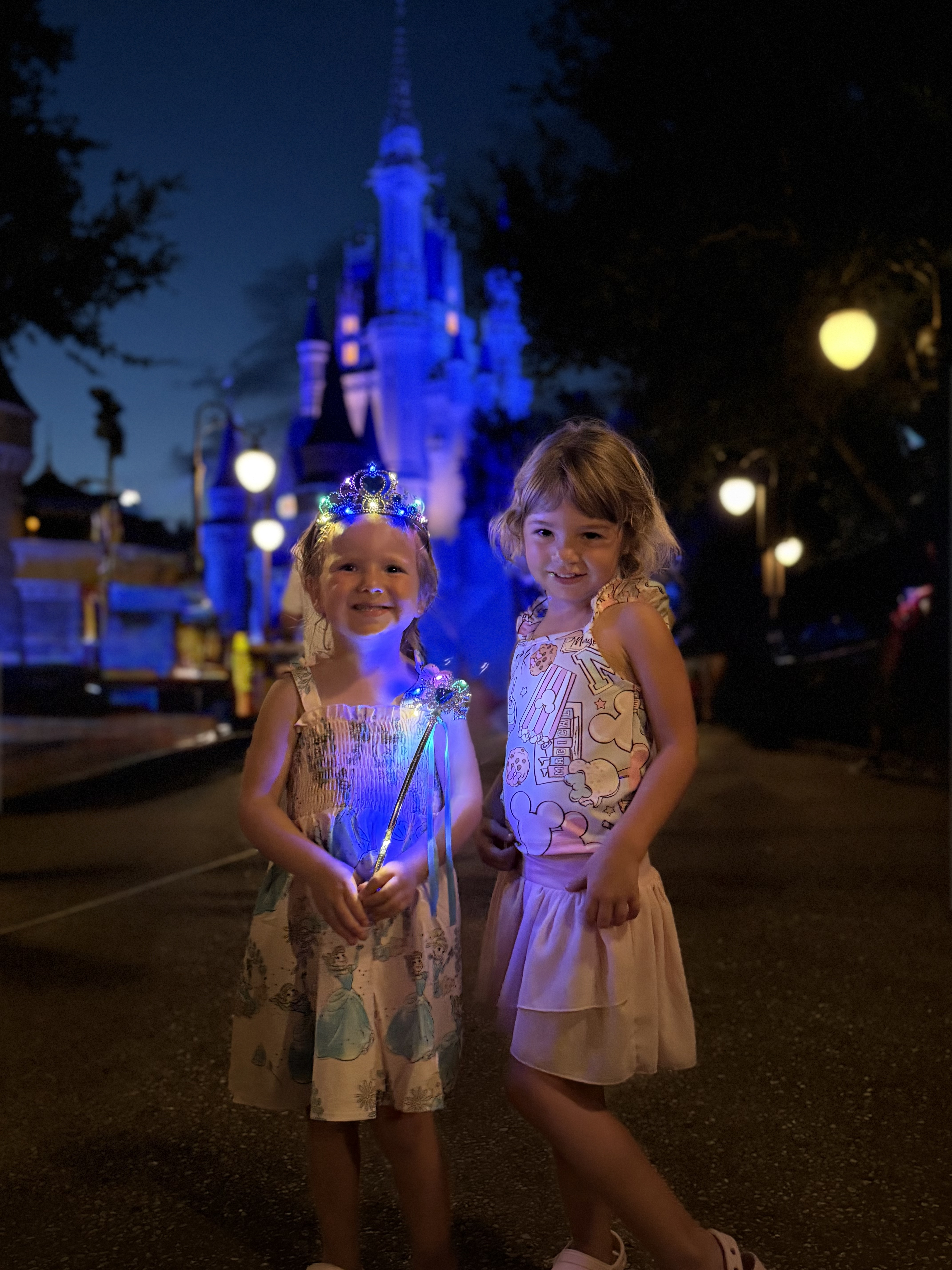 Sarah and Courtney's daughters in front of the castle at night, one wearing a light-up tiara and holding a glowing wand